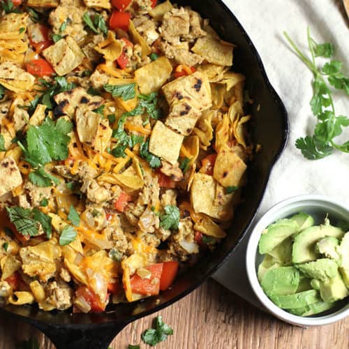 Overhead shot of a cast iron skillet of Migas, on a wooden background with a bowl of avocado slices.