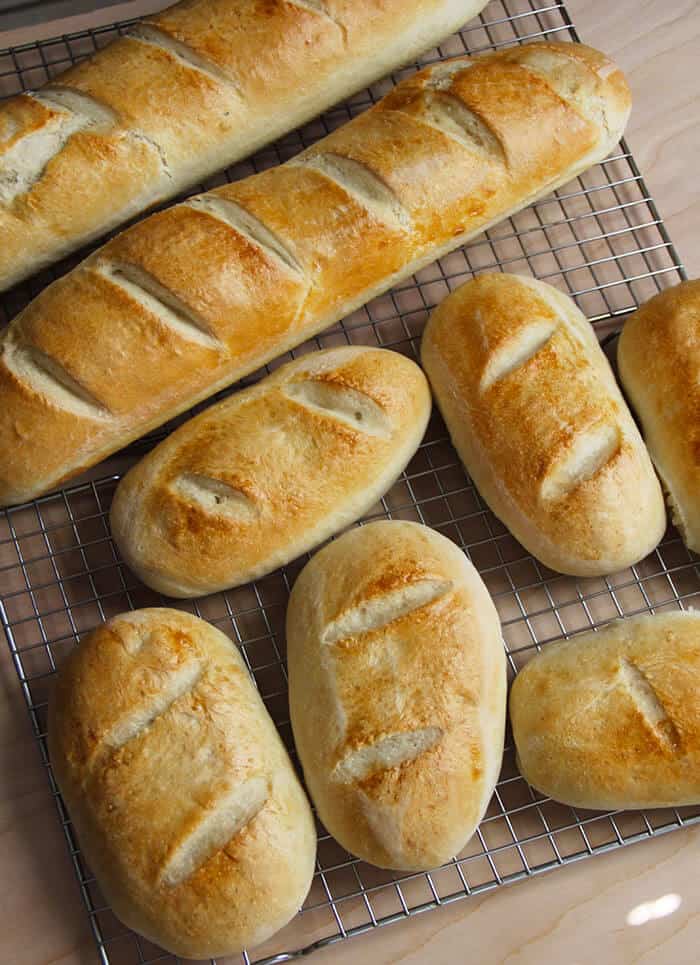 Overhead shot of two French bread loafs and several smaller loafs, all with slits cut on top and on a wire baking sheet.
