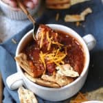 Overhead shot of a hand spooning a bite of slow cooker chili, in a white bowl on a gray tray with a blue napkin.