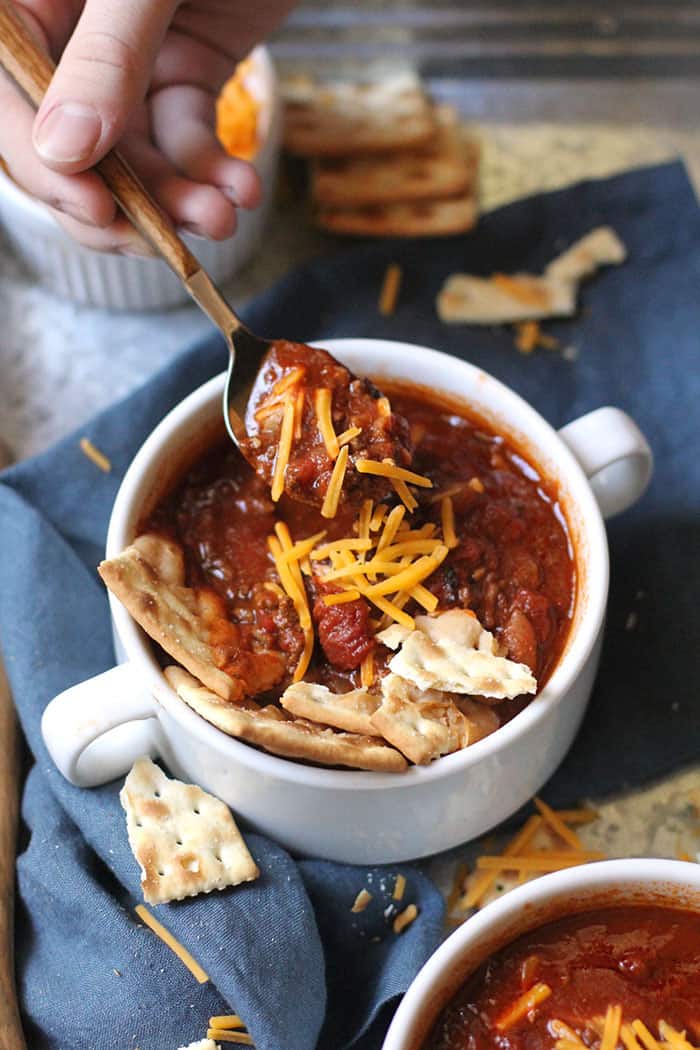 Overhead shot of a hand spooning a bite of slow cooker chili, in a white bowl on a gray tray with a blue napkin.