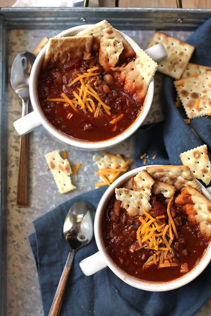 Overhead shot of two bowls of slow cooker chili, with crackers inside the bowls, on a gray tray with a blue napkin and spoons.