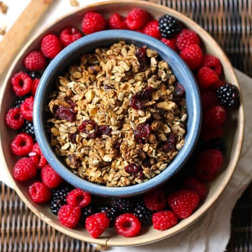 Overhead shot of a round blue bowl of granola, wet inside a tan shallow bowl, with raspberries and blackberries inside that bowl.