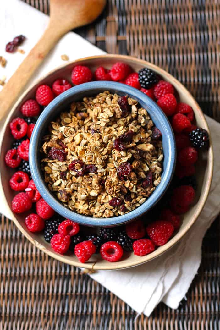 Overhead shot of a round blue bowl of granola, wet inside a tan shallow bowl, with raspberries and blackberries inside that bowl.