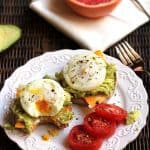 Two poached egg avocado muffins on a white plate, with a fork and a grapefruit half.
