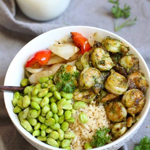 A white bowl filled with pesto shrimp, rice, peppers and onions, and edamame, on a gray napkin.