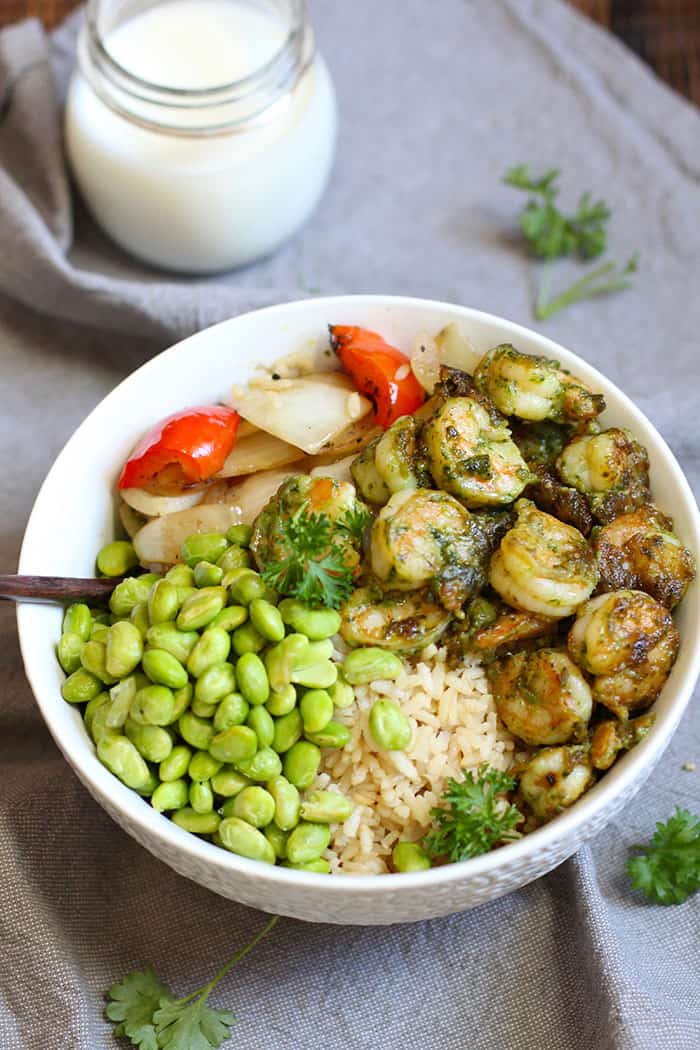 A white bowl filled with pesto shrimp, rice, peppers and onions, and edamame, on a gray napkin.