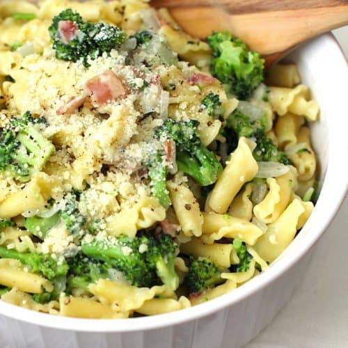 A closeup shot of a large white bowl of broccoli pasta with bacon, with a wooden spoon.