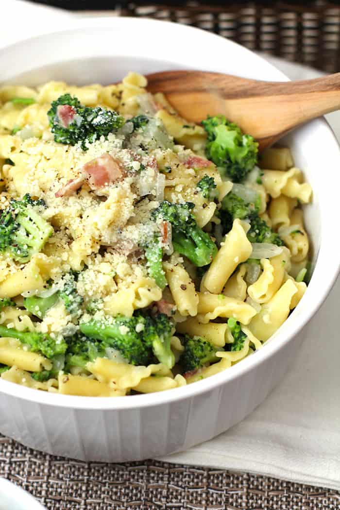 A closeup shot of a large white bowl of broccoli pasta with bacon, with a wooden spoon.