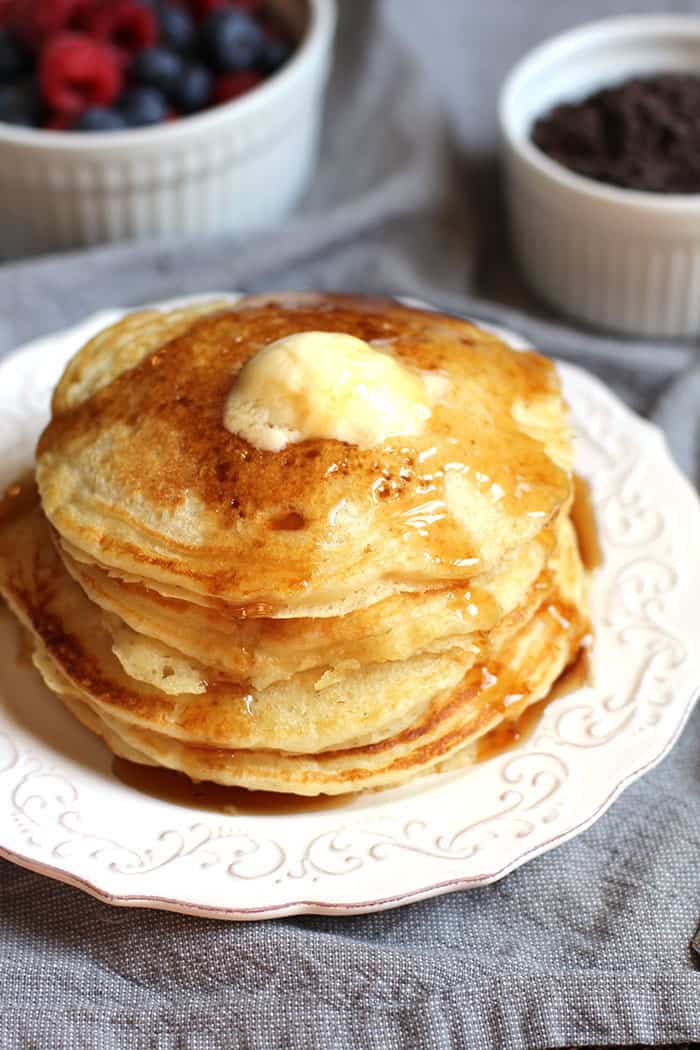 A large stack of buttermilk pancakes on a white plate with butter and syrup on top.