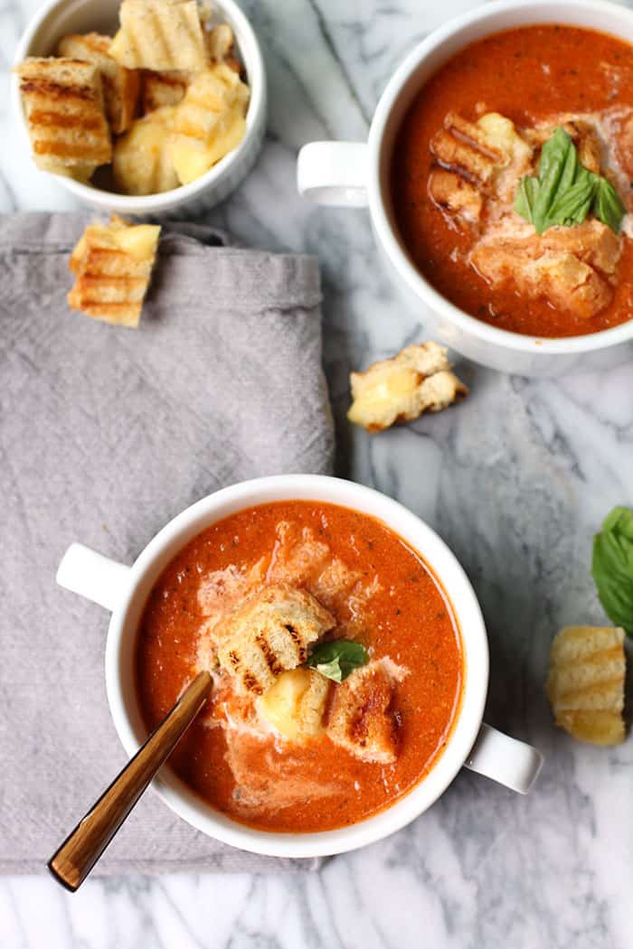 Overhead shot of two white bowls of tomato soup with grilled cheese croutons, all on a gray background.