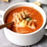 Overhead shot of two white bowls of tomato soup with grilled cheese croutons, all on a gray background.