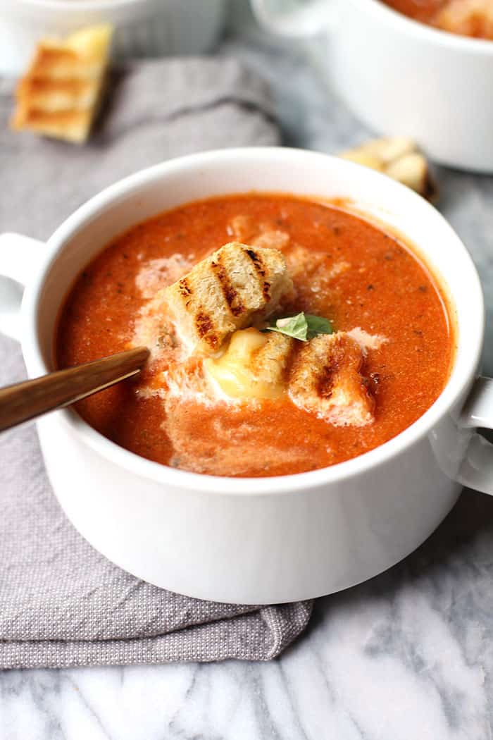 Overhead shot of a white bowl of tomato soup with grilled cheese croutons, on a gray background.