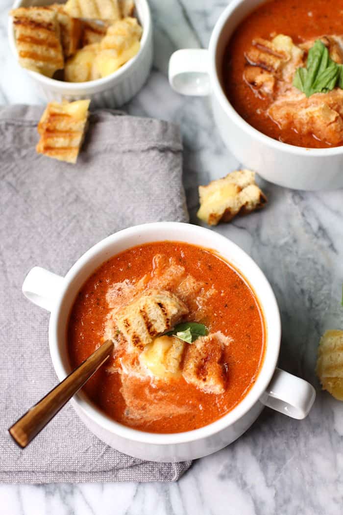 Overhead shot of two white bowls of tomato soup with grilled cheese croutons, all on a gray background.