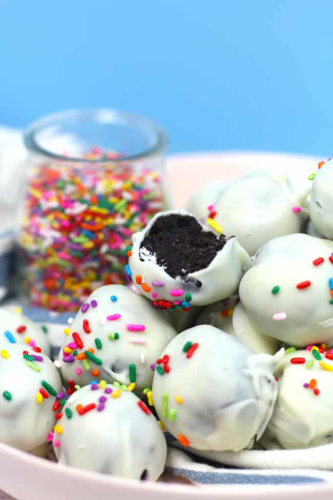 Side shot of a pink bowl of white chocolate Oreo Truffles, with one missing a bite, and a bowl of sprinkles, with a blue background.