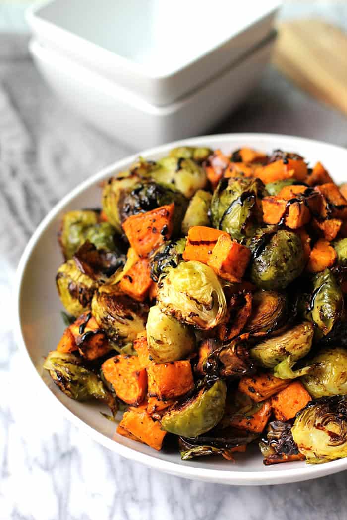 Side view of a heaping bowl of Brussels sprouts and sweet potatoes, with a balsamic drizzle on top, all in a white bowl on a gray and white background.