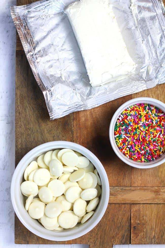 Overhead shot of a wooden board with a bowl of white choc melting wafers, a bowl of sprinkles, and a block of cream cheese.