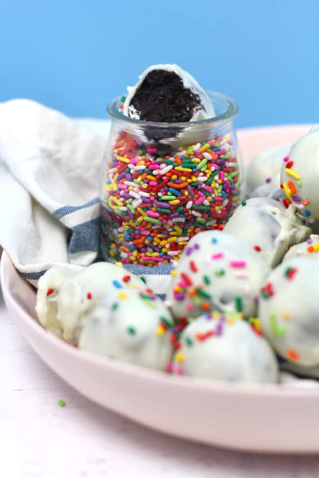 Side shot of a pink bowl of white chocolate Oreo Truffles, with a bowl of sprinkles and a partially eaten truffle inside it.