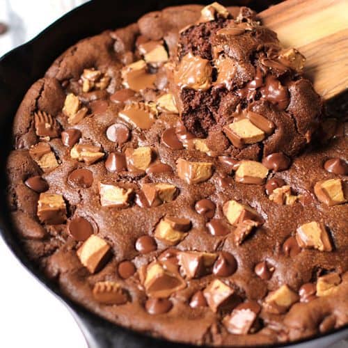 Side shot of a large skillet chocolate peanut butter cookie, with a wooden spoon dipping into the gooey baked cookie.