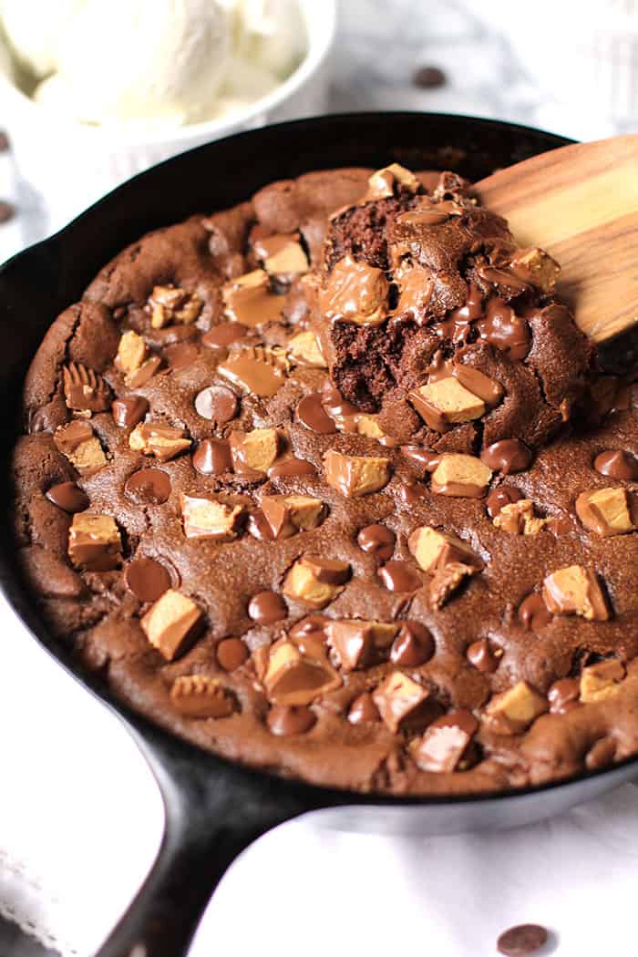 Side shot of a large skillet chocolate peanut butter cookie, with a wooden spoon dipping into the gooey baked cookie.