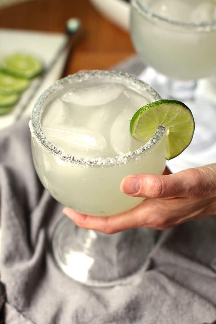 Overhead shot of a large glass of skinny margaritas, with a lime wedge, and a hand holding it, on a gray napkin.