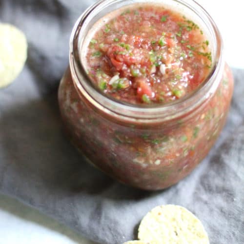 Shot of a mason jar of homemade salsa, on a gray napkin with a few chips beside it.