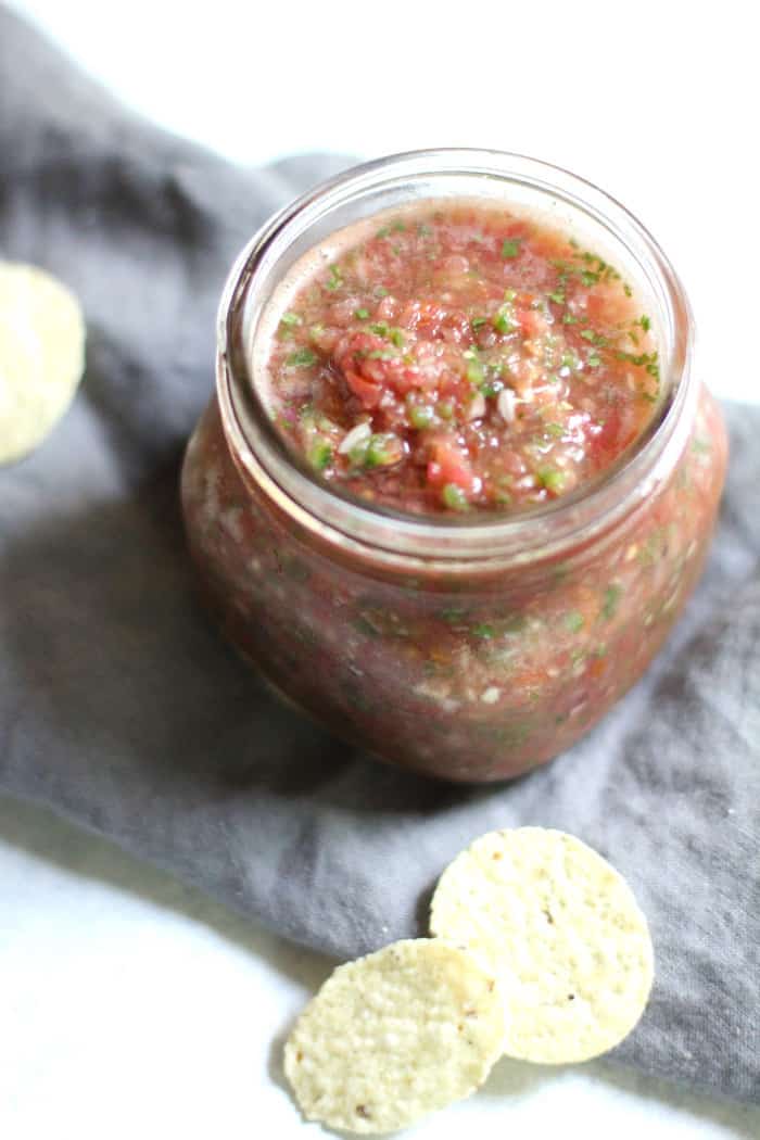 Shot of a mason jar of homemade salsa, on a gray napkin with a few chips beside it.