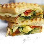 Side shot of a stack of two roasted vegetable pesto paninis, on a white background.