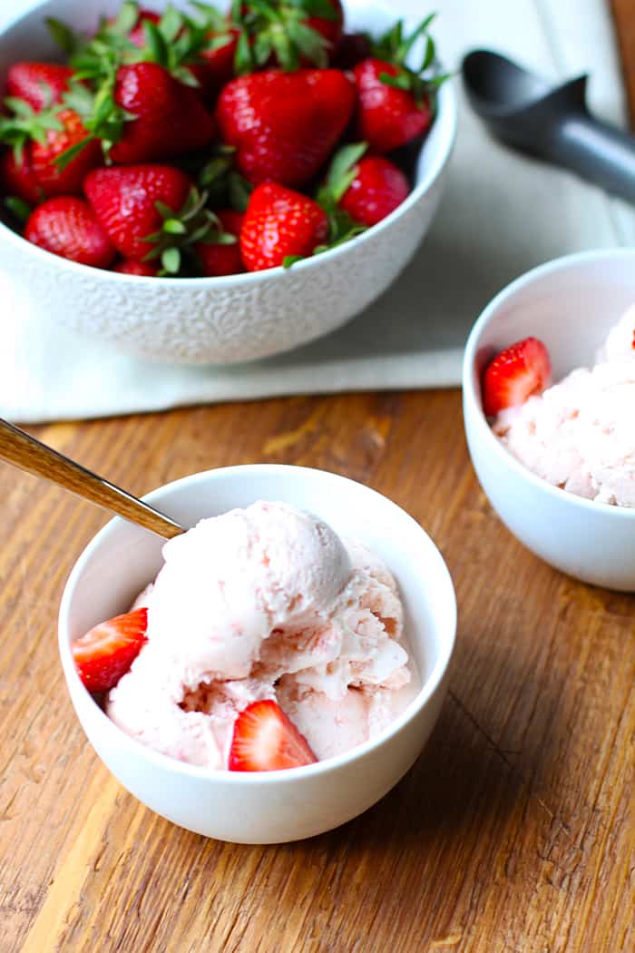 Overhead shot of two white bowls of homemade strawberry ice cream, on a wooden board, with a large bowl of whole strawberries in the background.
