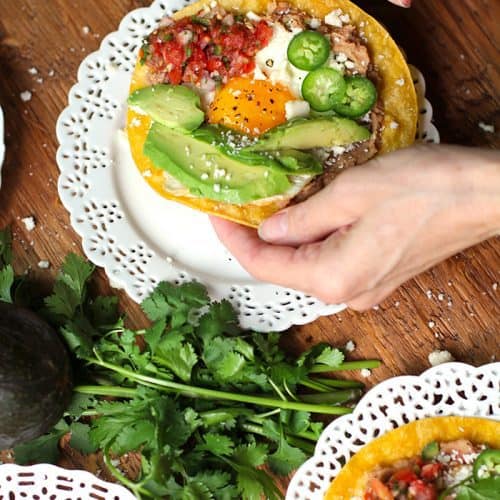 Two hands picking up a Huevos Rancheros Tostada, over a white plate.