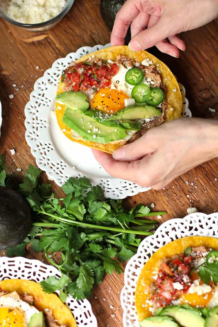 Two hands picking up a Huevos Rancheros Tostada, over a white plate.