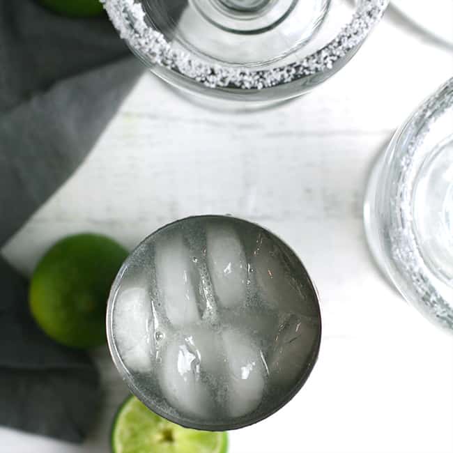 Overhead shot of a cocktail shaker filled with margarita, and some prepped margarita glasses.