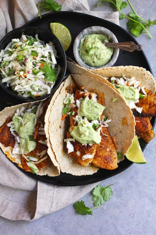 Foto in primo piano di tre facili tacos di pesce su un piatto nero, con insalata croccante e crema di avocado, su uno sfondo grigio.