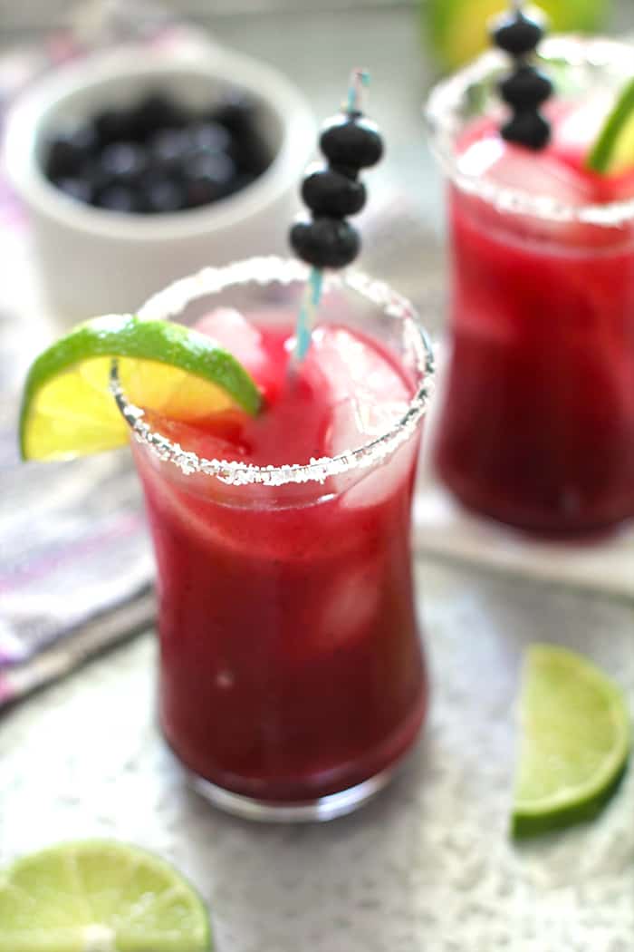 Two blueberry lime margaritas on a gray tray with lime wedges.