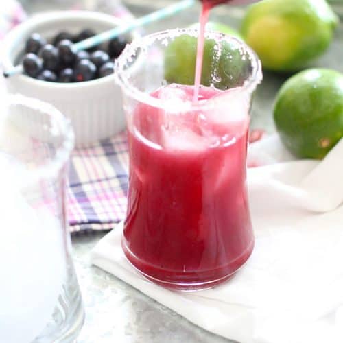 A cocktail shaker pouring a blueberry lime margarita into a glass with ice, on a tray.