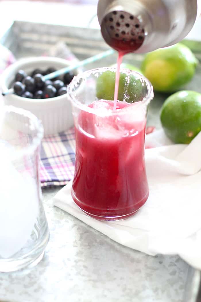 A cocktail shaker pouring a blueberry lime margarita into a glass with ice, on a tray.