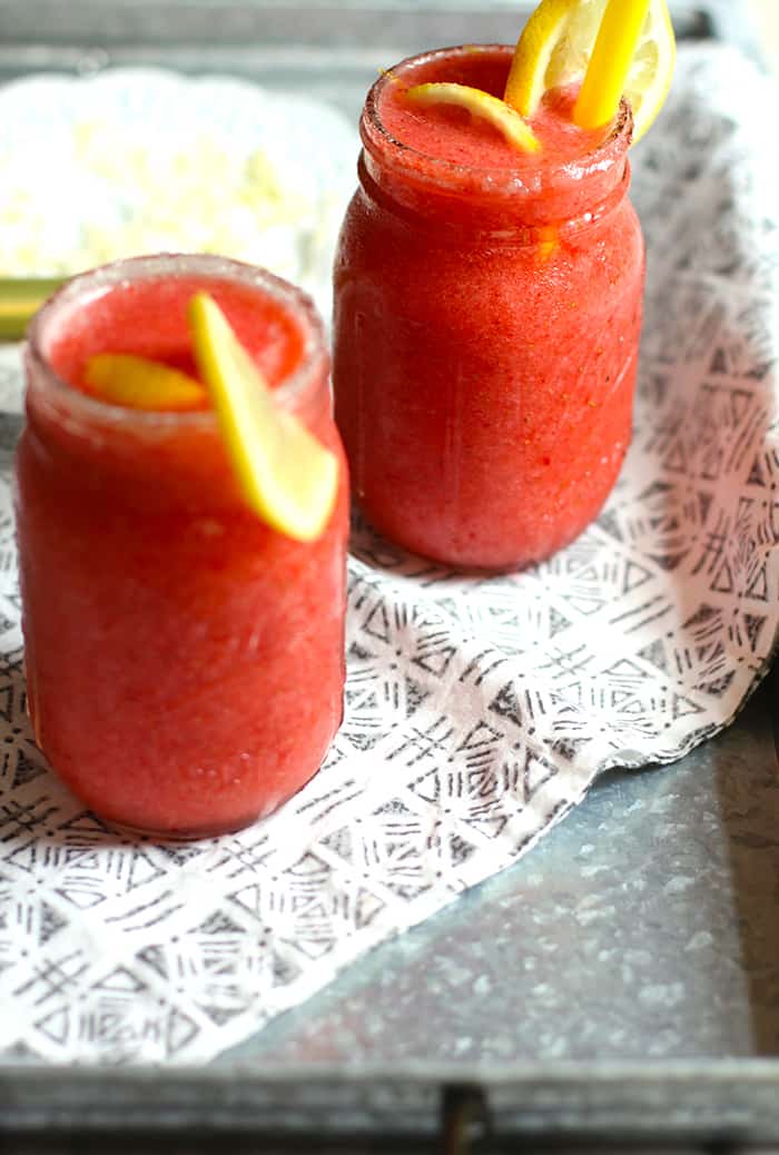 Side shot of my strawberry lemonade vodka slushies, in glass mason jars, with lemon garnish, on a gray tray.