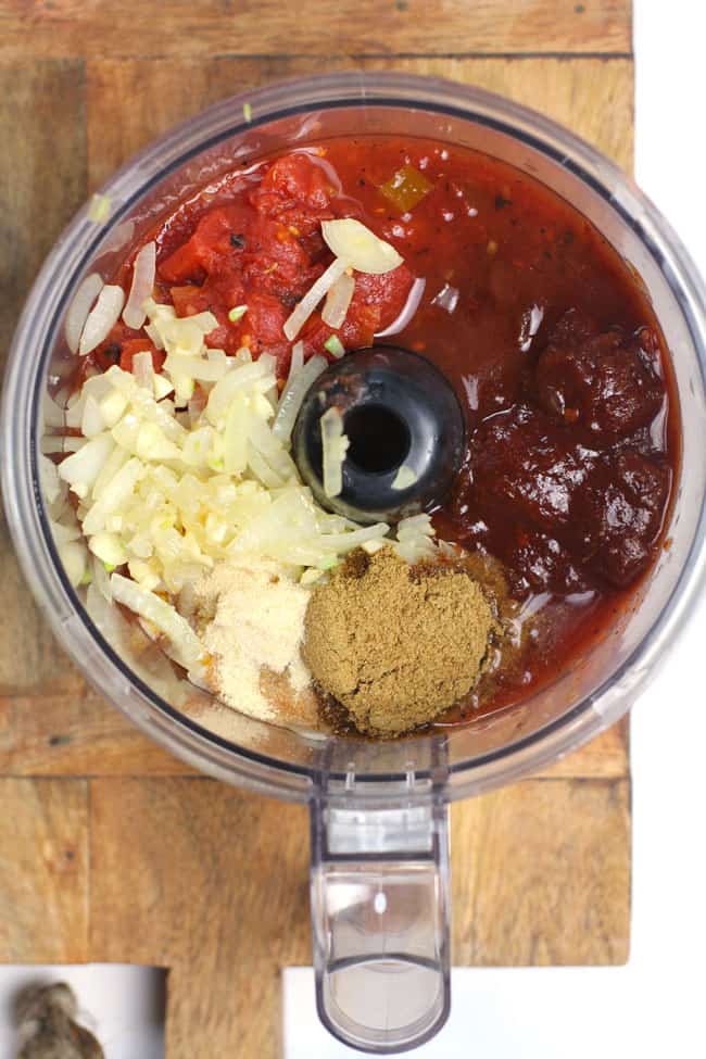 Overhead view of a food processor with sauce ingredients, on a wooden board.