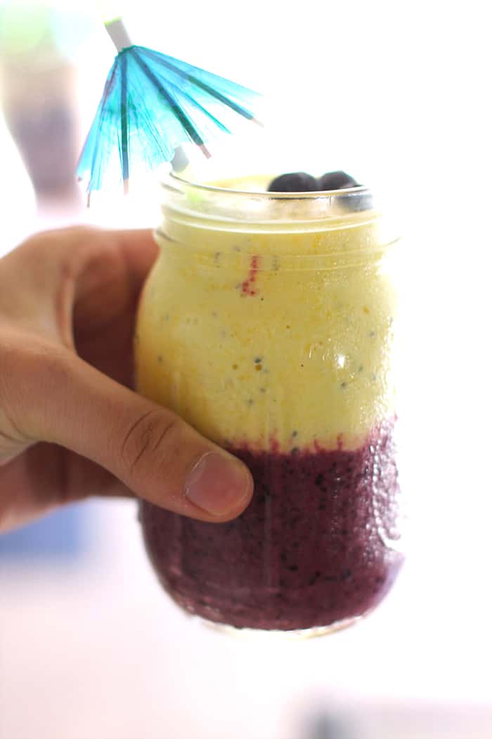 Side shot of a hand holding a blueberry mango chia smoothie in a mason jar, against a window.