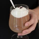 Side shot of a hand holding a dessert glass of creamy chocolate mousse, with a spoon inside.