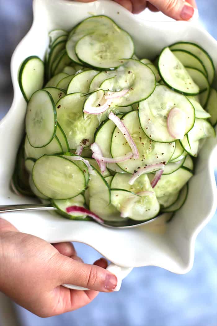 Two hands holding a white bowl of fresh cucumbers.