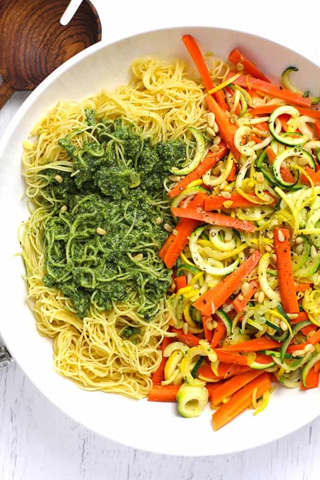 Overhead shot of a large white bowl of pesto vegetable pasta, on a white background.