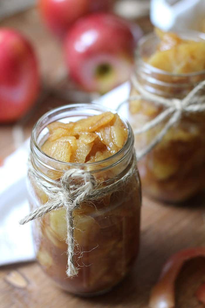 A mason jar with brown twine tied on it, full of homemade applesauce, on a brown background with bright red apples in the background and another jar of applesauce.