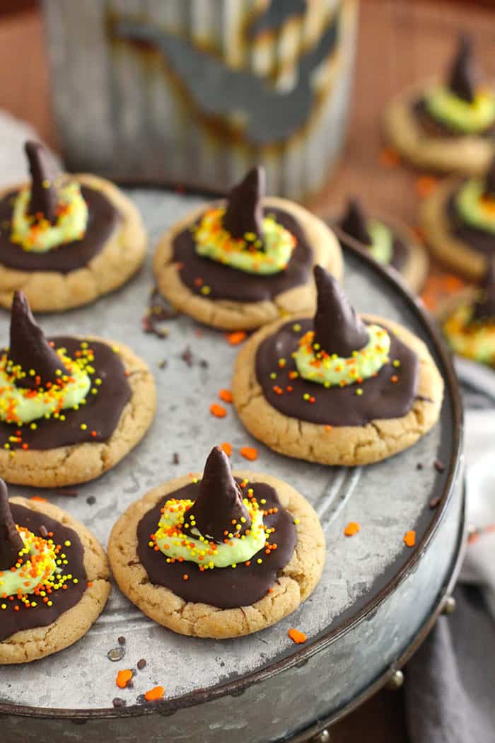 Side angle of a gray cake platter with witch hat peanut butter cookies on top, with a halloween decoration in the background and more cookies.