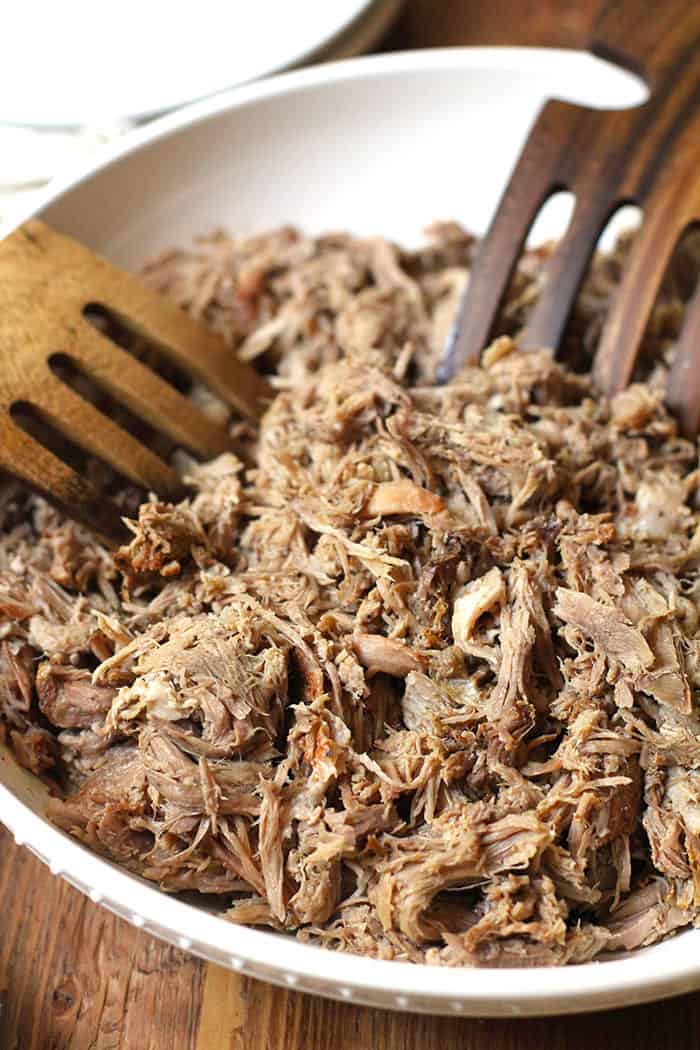 Overhead close-up shot of a large white bowl with shredded pulled pork and two large wooden forks inside it.