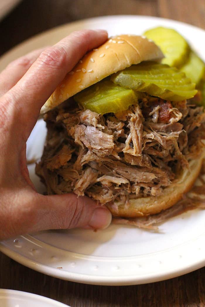 Side shot of my hand grabbing a very large pulled pork sandwich with pickles, on a white plate.