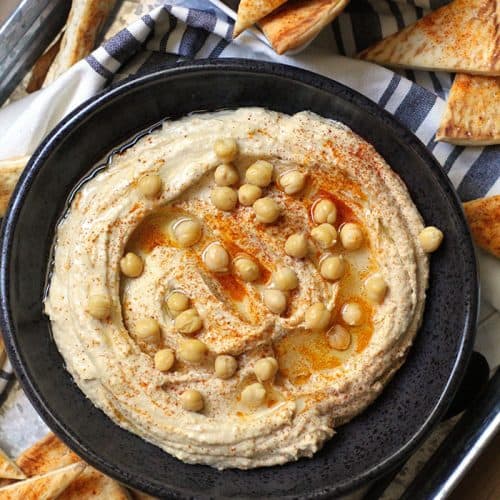 Overhead shot of a black bowl of homemade hummus with baked pita chips beside it on a gray tray.