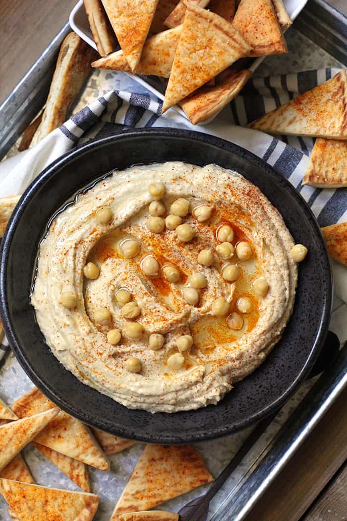 Overhead shot of a black bowl of homemade hummus with baked pita chips beside it on a gray tray.