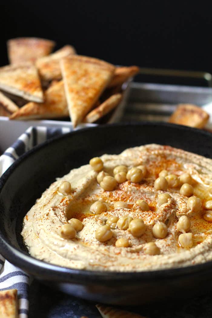 Side view of a black bowl of homemade hummus with baked pita chips beside it on a gray tray.