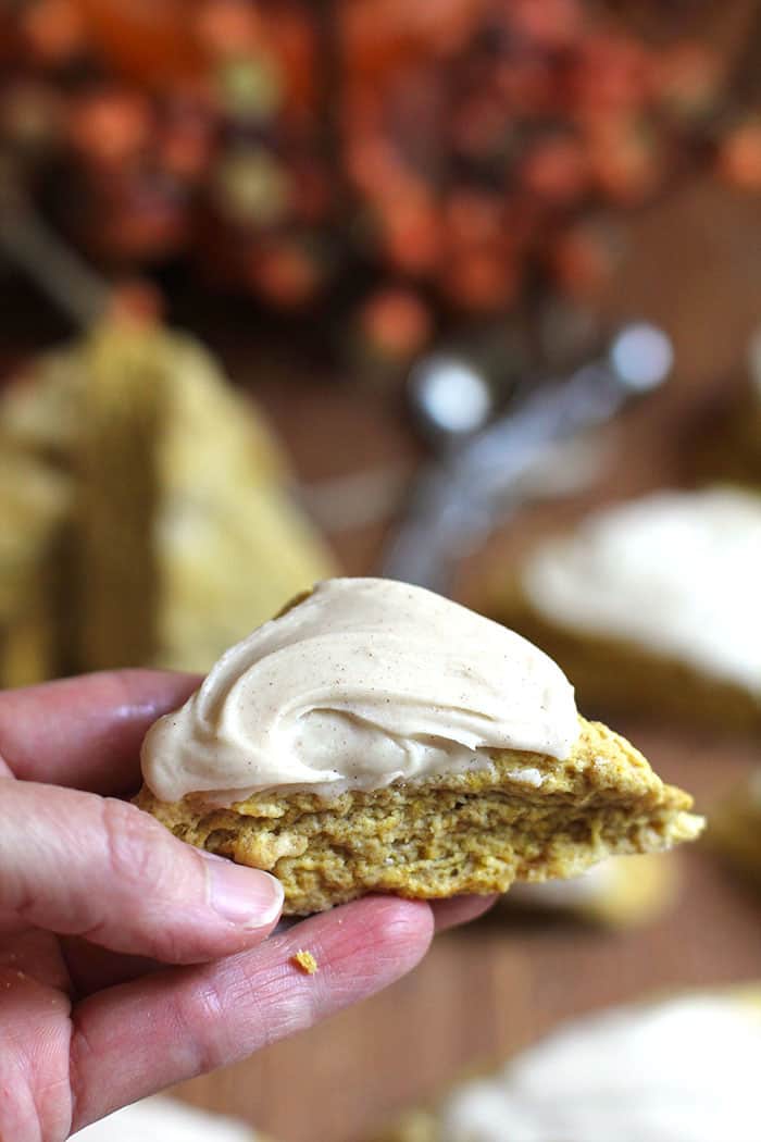 A side shot of my hand holding a mini iced pumpkin scone, with some pumpkin decorations in the background.