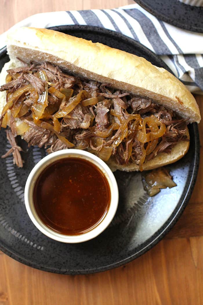 Overhead shot of beef au jus sandwich on a black plate, with a small white bowl of au jus, on a wooden background.
